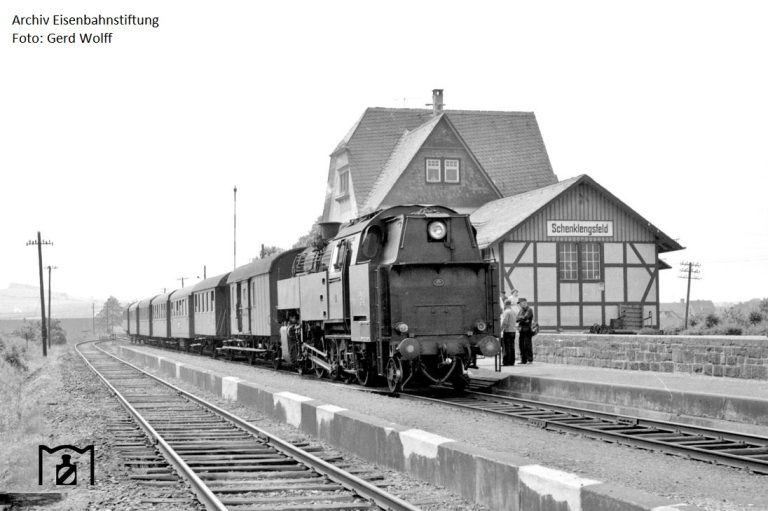 Schwarz-Weiß-Foto eines Zuges an einem Bahnhof mit einem historischen Gebäude.
